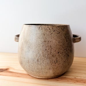 May include: A rounded ceramic pot with a speckled beige and brown finish, featuring two small side handles and a wide opening. The vessel has a textured surface with darker brown flecks distributed across the light tan body.