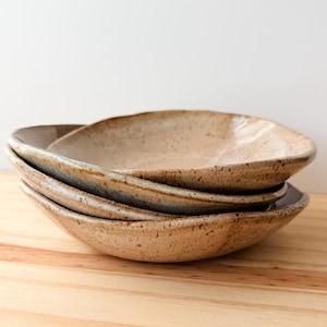 May include: A stack of three ceramic bowls with an irregular, organic shape. The bowls feature a speckled beige and tan glaze with subtle blue-grey undertones along the rims. The surfaces have a textured, earthy appearance with visible dark brown flecks.