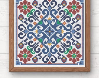 May include: A framed cross-stitch pattern featuring a symmetrical design with blue, red, green, and yellow floral and geometric elements. The artwork is set against a white background and framed in a light brown wooden frame. The PDF logo is visible.