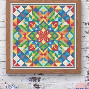 Op de afbeelding: Een ingelijst kruissteekpatroon met een geometrisch ontwerp. Het kunstwerk toont een symmetrische opstelling van kleurrijke driehoeken, vierkanten en ruiten in tinten rood, geel, blauw en groen. Het frame is van hout en hangt aan een witte bakstenen muur.