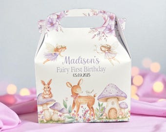 May include: A white party favor box with a handle, decorated with fairies, flowers, mushrooms, and woodland creatures. The text "Madison's Fairy First Birthday 05.09.2025" is printed on the front. The box is on a pink surface.