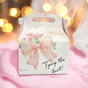 Pode incluir: Caixa de presente branca com um laço rosa e um design floral. A caixa tem uma alça e diz "Tying the knot!" na frente.
