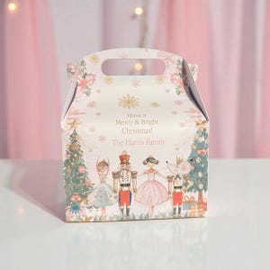 Puede incluir: Una caja de regalo navideña blanca con asa, decorada con copos de nieve y detalles florales. La caja presenta una ilustración festiva de personajes del ballet El Cascanueces, con el texto "Have a Merry & Bright Christmas! The Harris Family."