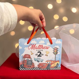 Op de afbeelding: Een rechthoekige papieren cadeauzak met rode linthenvaten en een blauwe luchtachtergrond met wolken. De zak toont de tekst 'Matthew is turning 2' naast illustraties van een rode vrachtwagen, een blauwe auto, een rode raceauto, een trofee en een verkeerslicht.