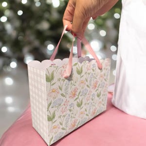 以下が含まれることがあります： 花柄とピンクのリボンハンドルが付いた小さなギフトバッグ。バッグはスカラップエッジと市松模様の側面が特徴です。フローラルデザインには、ピンク、青、緑の花が含まれています。