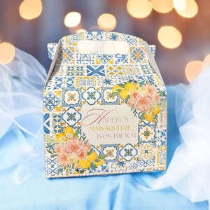 Pode incluir: Uma caixa de presente branca com um padr&atilde;o de azulejos azuis e amarelos e detalhes florais. A caixa tem uma al&ccedil;a e diz "Hadeel's Main Squeeze is on the way".