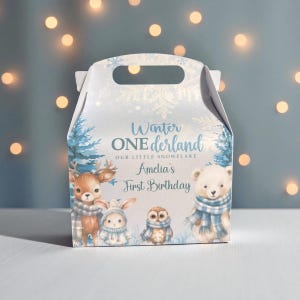 Op de afbeelding: Een lichtblauwe geschenkdoos met een handvat, met winterbosdieren en de tekst "Winter ONEderland" en "Amelia's First Birthday". De doos heeft illustraties van een hert, een konijn, een uil en een ijsbeer.