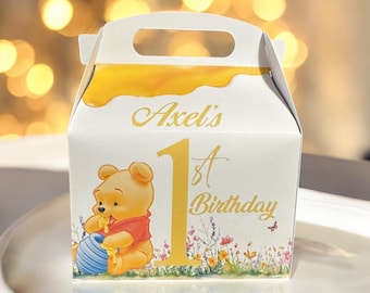 May include: A Winnie the Pooh themed party favor box with a yellow honey drip design. The box is white with gold accents and features a picture of Winnie the Pooh holding a honey pot. The text on the box reads "Axel's 1st Birthday".