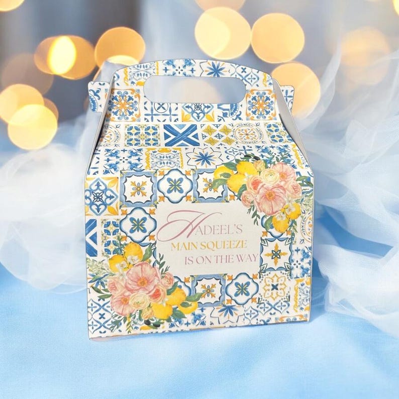 Pode incluir: Uma caixa de presente azul e branca com padr&atilde;o e al&ccedil;a. A caixa tem um design floral com lim&otilde;es e o texto "Hadeel's Main Squeeze is on the way".