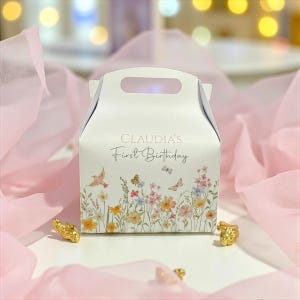 Puede incluir: Una caja de regalo blanca con asa, con el texto "CLAUDIA'S First Birthday". La caja está decorada con un diseño floral de mariposas, pájaros y flores silvestres en colores pastel. Piezas decorativas doradas se colocan alrededor de la caja.