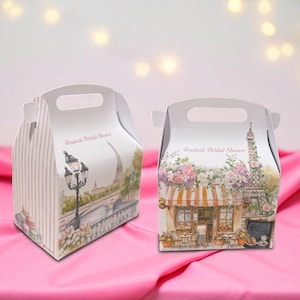 Puede incluir: Dos cajas blancas con forma de cabaña con asas, con escenas parisinas. Una caja tiene una ilustración de farola y puente, la otra un café y la Torre Eiffel. El texto en ambas cajas dice "Jessica's Bridal Shower."