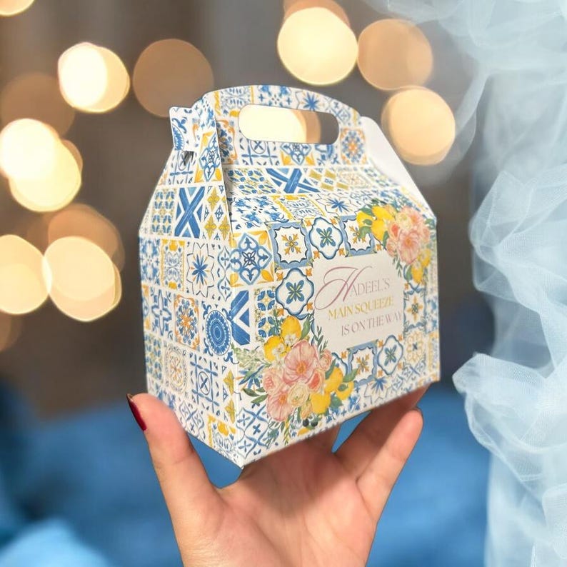 Pode incluir: Uma caixa de presente com padr&atilde;o branco, azul e amarelo com al&ccedil;a. A caixa tem um design floral e o texto "Adeel's Main Squeeze is on the way".