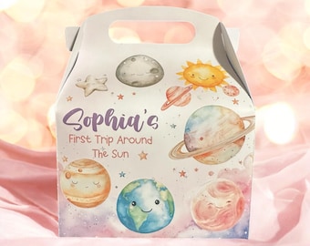 May include: A white party favor box with a handle, decorated with watercolor illustrations of planets, stars, and a smiling sun. The text "Sophia's First Trip Around The Sun" is written in purple script.