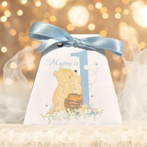 Op de afbeelding: Een witte cadeaubox met een blauw lint, waarop Winnie de Poeh met een honingpot is afgebeeld. De doos heeft de tekst "Mateo is 1" erop gedrukt.