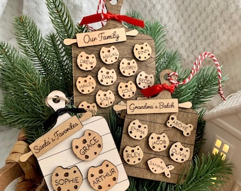 May include: Wooden Christmas tree decorations in the shape of cutting boards, with cookie-shaped name tags. One says "Our Family," another "Grandma's Batch," and the third "Santa's Favorites." Each has a rolling pin accent and a red and white string for hanging.