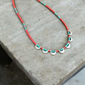 Könnte beinhalten: Halskette mit roter Kordel, türkisen und silbernen Details. Die Halskette hat silberne, halbmondförmige Anhänger mit türkisen Inlays. Das Design ist schlicht und elegant, passend für den Alltag.