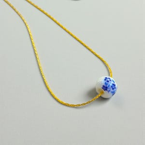 K&ouml;nnte beinhalten: Eine zarte, goldfarbene Halskette mit einem wei&szlig;en Keramikperlenanh&auml;nger. Die Perle ist mit einem blauen Blumenmuster verziert. Die Halskette wird vor einem hellgrauen Hintergrund pr&auml;sentiert.