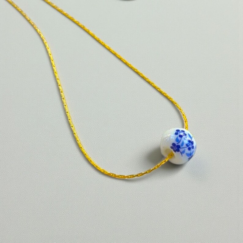 K&ouml;nnte beinhalten: Eine zarte, goldfarbene Halskette mit einem wei&szlig;en Keramikperlenanh&auml;nger. Die Perle ist mit einem blauen Blumenmuster verziert. Die Halskette wird vor einem hellgrauen Hintergrund pr&auml;sentiert.