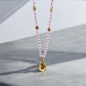 Könnte beinhalten: Eine lange Halskette mit einem goldenen Anhänger und weißen Perlen. Die Halskette hat eine rote Schnur und goldene Scheibenakzente. Der Anhänger hat eine strukturierte, unregelmäßige Form. Die Halskette wird vor einem grau-weißen Hintergrund präsentiert.