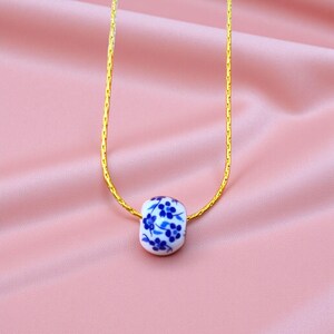 K&ouml;nnte beinhalten: Goldfarbene Halskette mit einem wei&szlig;en Keramikanh&auml;nger. Der Anh&auml;nger ist mit einem blauen Blumenmuster verziert. Die Halskette wird auf einem rosa Satin-Hintergrund pr&auml;sentiert. Der Anh&auml;nger ist ca. 1,5 cm lang.