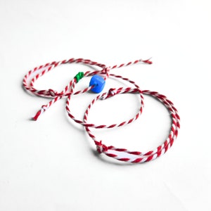 Pode incluir: Três pulseiras de corda vermelha e branca amarradas juntas com uma conta azul no centro.