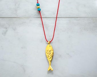 May include: Gold-colored fish pendant necklace on a red cord. The fish pendant has a textured surface and a small gemstone. The necklace also features turquoise and gold-colored beads.