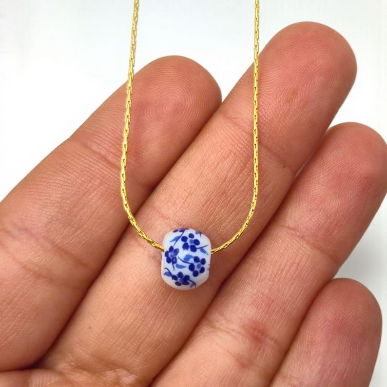 K&ouml;nnte beinhalten: Eine zarte goldfarbene Halskette mit einer wei&szlig;en Keramikperle. Die Perle ist mit einem blauen Blumenmuster verziert. Die Halskette wird vor einem neutralen Hintergrund pr&auml;sentiert.
