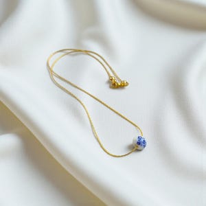 K&ouml;nnte beinhalten: Zarte goldfarbene Halskette mit einem einzelnen, runden Anh&auml;nger. Der Anh&auml;nger ist wei&szlig; mit einem blauen Blumenmuster. Die Halskette liegt auf einem wei&szlig;en, strukturierten Stoff und erzeugt eine schlichte und elegante &Auml;sthetik.