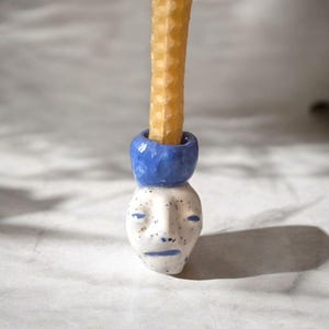 May include: A ceramic candle holder with a face design. The holder is white with blue accents and small black speckles. A yellow candle is inserted into the blue top of the holder. The candle holder has a unique, artistic design.