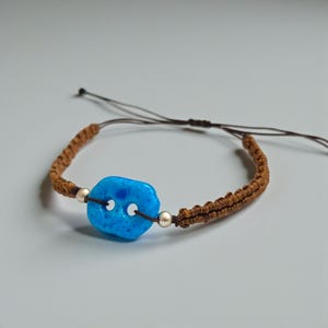 May include: A handmade macrame bracelet featuring a vibrant blue stone centerpiece. The bracelet has a brown woven band and is accented with silver-colored beads. The adjustable cord allows for a comfortable fit. This unique bracelet is a stylish accessory.