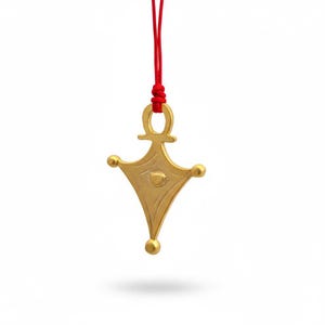 Könnte beinhalten: Goldfarbene Anhänger-Halskette mit geometrischem Design, an einer roten Kordel befestigt. Der Anhänger hat eine rautenförmige Form mit abgerundeten Kanten, verziert mit drei kleinen Kugeln und einem augenähnlichen Detail.