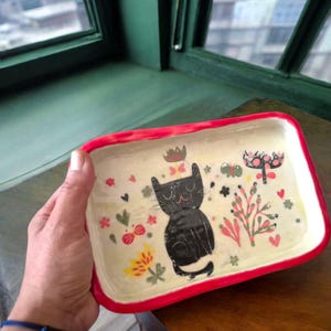 May include: A rectangular ceramic tray with a red rim. The tray features a black cat illustration surrounded by colorful floral and heart designs. The background is off-white.