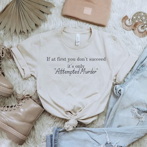 May include: A cream-colored t-shirt with the text "If at first you don't succeed it's only 'Attempted Murder'" in black. The shirt is styled with light-wash denim jeans, tan boots, and a beanie. The items are arranged on a white, fluffy surface.