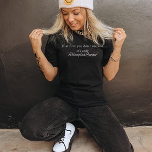 May include: A woman wearing a black t-shirt with the text "If at first you don't succeed, it's only Attempted Murder."  She is wearing a white beanie with a yellow smiley face on it and black jeans.