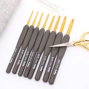 Peut inclure: Un ensemble de crochets gris avec des pointes dorées, de tailles allant de 2,00 mm à 6,00 mm, et une paire de ciseaux à poignée dorée. Les crochets sont marqués de leur taille et de la marque "Tulip".