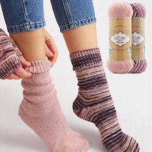May include: A pair of knitted socks, one solid light pink and the other with a striped pattern in shades of purple, pink, and beige. The word "alize" is visible in the upper left corner. Two spools of yarn are in the background.