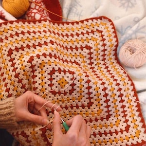 May include: A person is crocheting a large granny square blanket with beige, red, and yellow yarn. The blanket is laid out on a white surface.