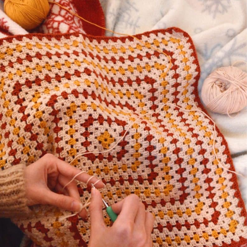 May include: A person is crocheting a large granny square blanket with beige, red, and yellow yarn. The blanket is laid out on a white surface.