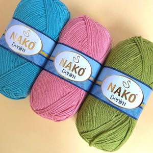 May include: Three balls of NAKO Denim yarn in teal, pink, and green. Each ball has a blue and gold label. The yarn is tightly wound, showing the texture of the fibres. The background is a light beige.