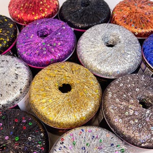 May include: A collection of colorful yarn skeins, each wrapped with shimmering sequins. The skeins are in various colors, including red, black, orange, purple, silver, gold, brown, and blue. The yarn is tightly wound, and the sequins catch the light.