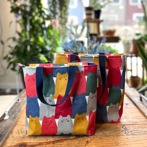 May include: Two colorful tote bags with a repeating pattern of cat faces in red, yellow, blue, gray, and green. The bags have navy blue handles and zippers. The bags are sitting on a wooden surface.