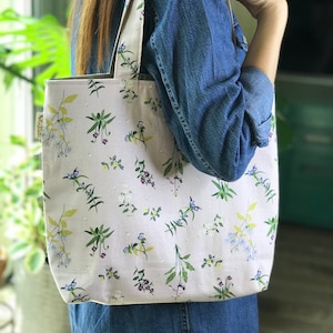 May include: A white tote bag with a floral pattern featuring small green leaves and purple, blue, and yellow flowers. The bag has two long straps.
