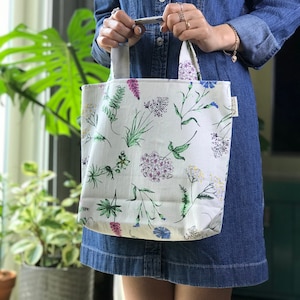 May include: A white tote bag with a floral print featuring green leaves, purple, pink, and blue flowers. The bag has two handles and a white label with black text.