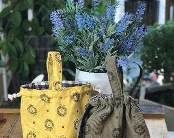 May include: Two small, corduroy fabric bags with drawstring closures. One is yellow with a black circular pattern, and the other is olive green with a similar pattern. Both have handles and sit on a wooden surface.