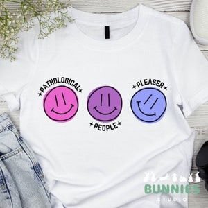 Könnte beinhalten: Weißes T-Shirt mit drei Smiley-Gesichtern. Das obere Smiley-Gesicht hat den Text "Pathological" darum herum. Das mittlere Smiley-Gesicht hat den Text "People" darunter. Das untere Smiley-Gesicht hat den Text "Pleaser" darum herum.