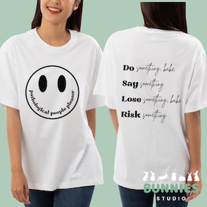 May include: White t-shirt with a black smiley face and the text "pathological people pleaser" on the front. The back of the shirt has the text "Do something, babe. Say something. Lose something, babe. Risk something." and a logo with the text "BUNNIES STUDIO".