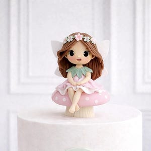 Könnte beinhalten: Eine skurrile Feenfigur mit braunen Haaren, einem Blumenkranz und weißen Flügeln. Sie trägt ein rosa Kleid und sitzt auf einem rosa Pilz mit weißen Punkten. Die Fee ist eine dekorative Tortenaufsatz.