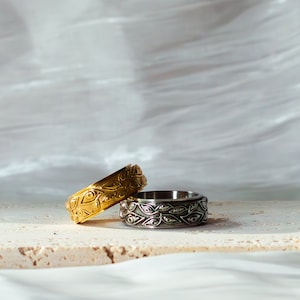 May include: Two rings with detailed patterns are displayed. One is gold-coloured, the other silver-coloured. Both rings have a repeating design of eye-shaped and leaf-like motifs. They are placed on a light-coloured surface.