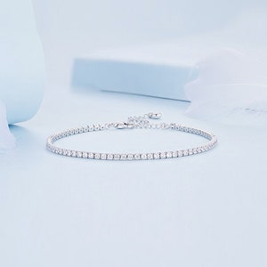 Op de afbeelding: Een zilveren armband met een rij kleine, sprankelende stenen. De armband is geplaatst tegen een lichtblauwe achtergrond, wat een strakke en elegante uitstraling creëert. De sluiting is zichtbaar en de armband lijkt verstelbaar.