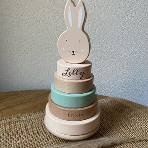 Op de afbeelding: Een houten stapelspel in de vorm van een konijn met een roze en witte kleurstelling. Het speelgoed heeft vier ringen, waarvan twee van natuurlijk hout zijn, één lichtgroen en één roze. De bovenste ring heeft de naam "Lilly" erop geschreven en de tweede ring vanaf de onderkant heeft de naam "Trixie" erop geschreven.